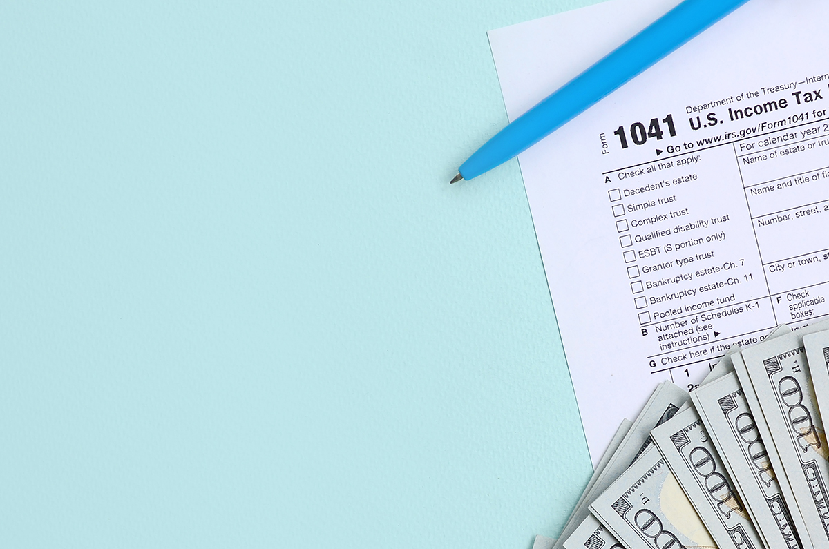 A blue pen and several hundred dollar bills are laying on top of a 1041 U.S. Income Tax form on a light blue background.